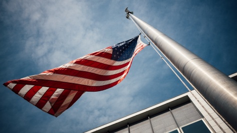 Looking up at a blue, slightly cloudy sky. A flagpole stretches almost straight up, with an American flag waving at the top.