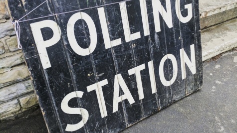A wooden sign painted black with large, white capital letters sitting on the ground outside a stone building reads, "POLLING STATION."
