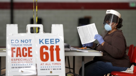 Image of a poll worker shifting through ballots, wearing a mask and gloves, in front of signs that say "keep 6ft distance"
