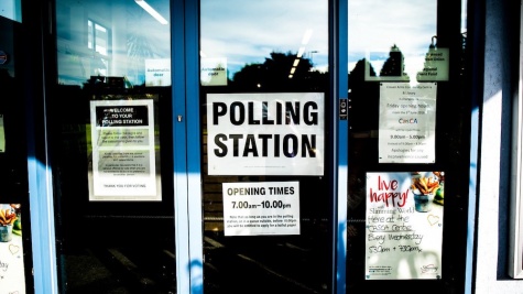 Automatic glass doors with a blue frame are plastered with several posters and welcome signs. A large white sign with black writing prominently reads "POLLING STATION" in black letters. The opening times are listed in a smaller sign below.