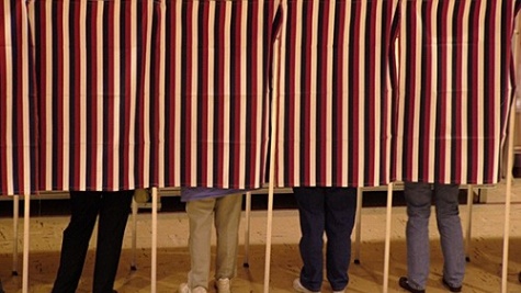 The legs of four people are seen side-by-side voting in adjacent voting booths.