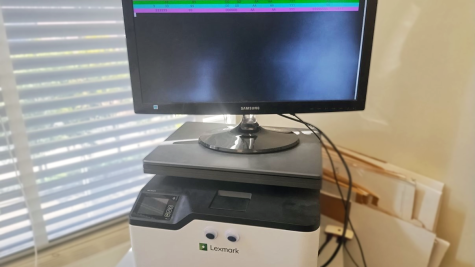 A computer monitor hooked up to a Raspberry Pi microcomputer shows a flashing rainbow display that says "39 days!" The monitor is perched atop a printer that has googly eyes on it, next to a window and in front of some folded cardboard boxes.