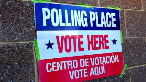 A brick wall with a sign taped to it with green masking tape. The sign is striped red, white, and blue; the text on the sign reads, "Polling place, vote here. Centro de votacion, vote aqui."