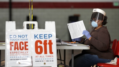 A Black woman sorts ballots or paperwork at a folding table. A folding sign in front of her provides instructions for social distancing. She is wearing a surgical mask and a face shield.