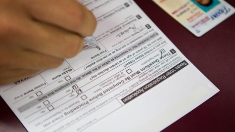 A Texas Voter registration application lays on a table; we can see a brown hand with a pen as someone fills it out. A Texas driver's license is off to one side.