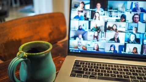A blue-green handmade ceramic mug sits on a dark wooden table next to a MacBook. On the screen, we can see a Zoom meeting full of people.