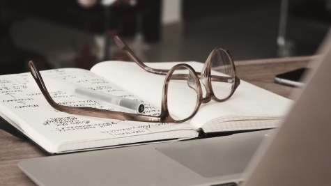 A pair of eyeglasses with thick brown translucent frames rests, upside down, on an open notebook. One page of the notebook is full of scribbled writing. A ballpoint pen rests next to one of the earpieces of the glasses.