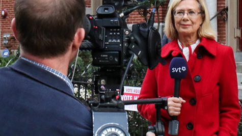 A woman in a red coat stands with a microphone in front of a building with a "vote here" sign. We are looking over the shoulder of a man in a suit coat with a TV camera at her.