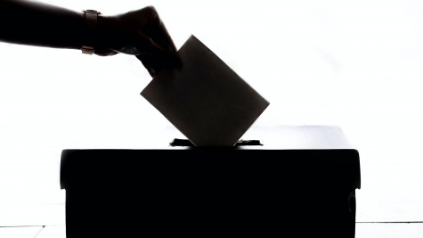 A backlit woman's hand is reaching out, placing a folded envelope into a ballot box.