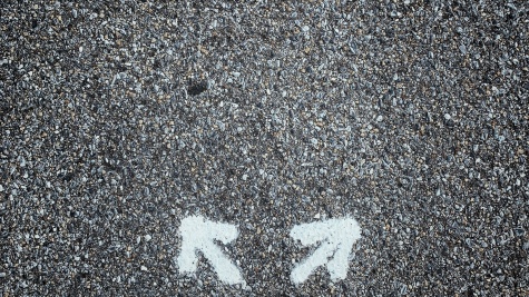 We are looking down as if at our own feet. The feet are wearing men's black dress shoes; the person is also wearing black slacks. Two small white arrows are spray painted on the asphalt in front of the person's feet.