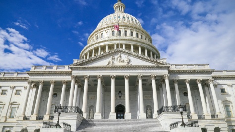 A view of the Capitol building. The camera is angled so that we are looking up the front steps at the dome, which stands against a bright blue sky.