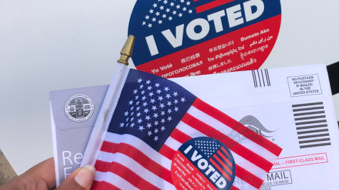 A close-up photo of a brown hand holding an American flag, a red white and blue "I voted" sticker, and a mail ballot envelope.