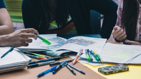 A table is covered in colored pens and markers. Three students are looking at notebooks and papers spread out in front of them. One student on the left is pointing to a point on a map in front of them with a highlighter.