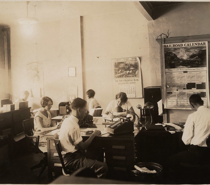 People are at work in the Missouri Pacific Railroad office in 1929. One man sits by a large adding machine.