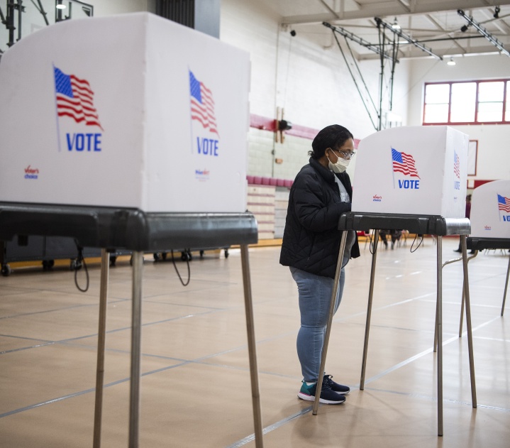 A woman of color, wearing a mask, jacket, and jeans, stands at a voting booth and fills out her ballot.