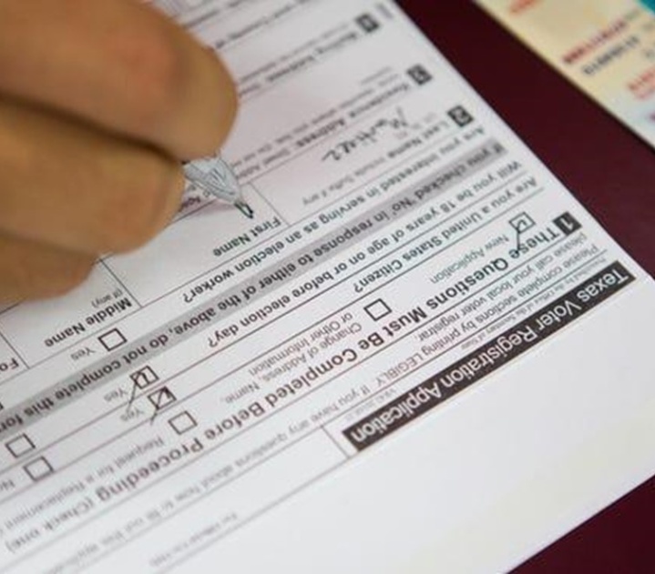 A Texas Voter registration application lays on a table; we can see a brown hand with a pen as someone fills it out. A Texas driver's license is off to one side.