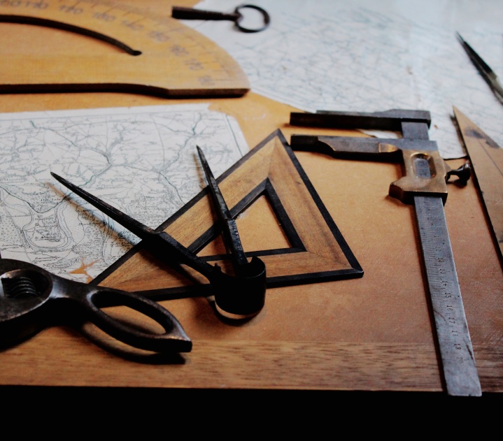 Mathematical tools, including a compass, a protractor, and a compass, lie on a wooden table on top of paper maps.