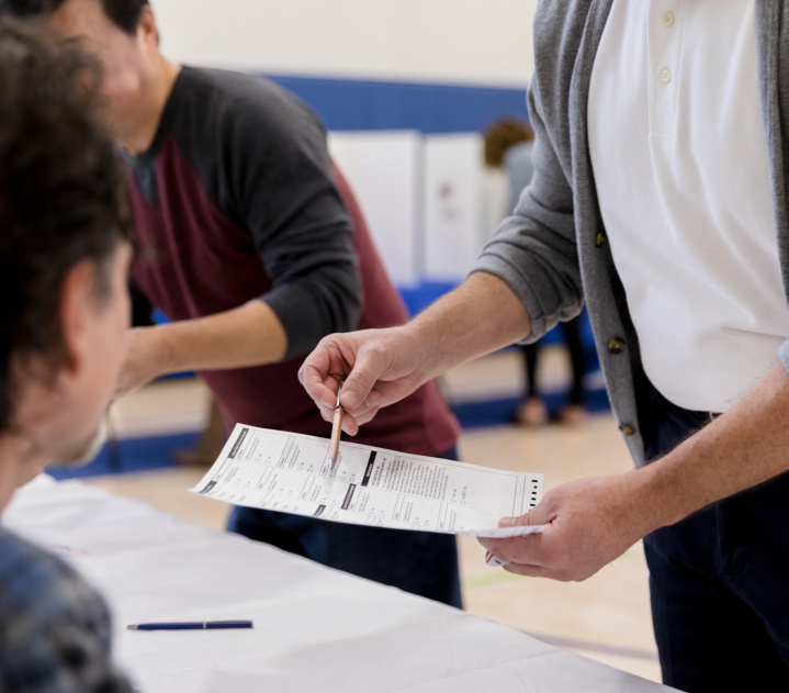 A person in a white shirt and unbuttoned grey cardigan uses a pen to point at a form in a polling place. They are standing in front of a table with a white table cloth, speaking with someone else who is seated just in front of the camera.