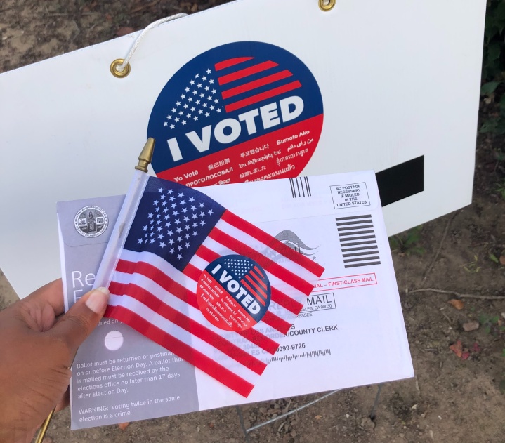 Someone holds an American flag with an "I voted" sticker on it, and an envelope for an absentee ballot