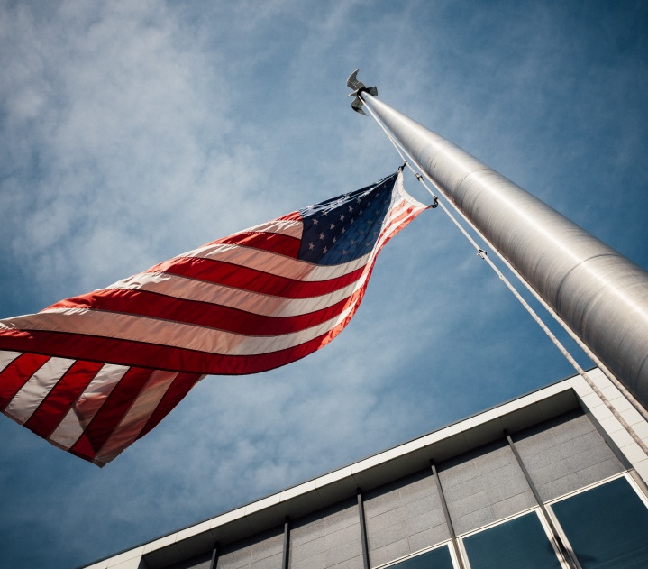 Looking up at a blue, slightly cloudy sky. A flagpole stretches almost straight up, with an American flag waving at the top.