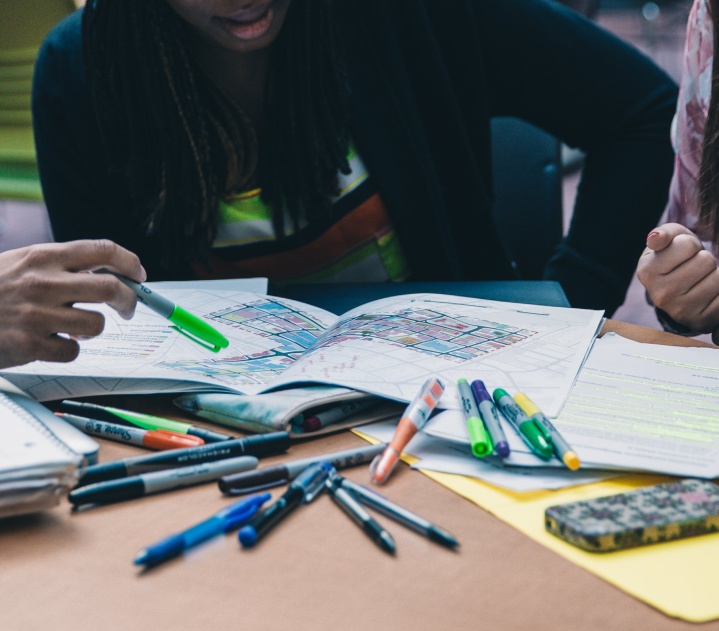 A table is covered in colored pens and markers. Three students are looking at notebooks and papers spread out in front of them. One student on the left is pointing to a point on a map in front of them with a highlighter.