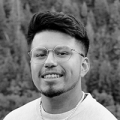 A young man with brown skin and black hair; he has rounded glasses, a short beard, and a wide smile.