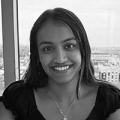 A young woman wearing a dark top and a necklace smiles at the camera. We can see the landscape of a small city out the large window behind her.