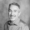 A man with short hair smiles at the camera; he is wearing a button-up shirt and is half-turned toward the viewer.
