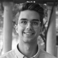 A young man wearing glasses and a collared shirt smiles at the camera. His hair is swept up away from his face; we can see two narrow columns and a garden arbor behind him.