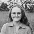 A black and white image of a young woman with long, slightly wavy hair and round glasses. She is smiling at the camera and wearing a collared shirt; behind her we can see a field and some distant trees.