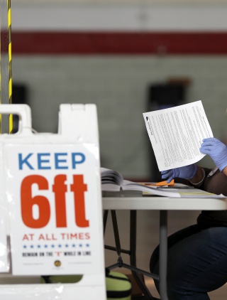 A black woman wearing a mask and face shield checks ballots. In front of the table she is seated at, there is a sandwich board sign with COVID health warnings, including requiring people to keep six feet apart.