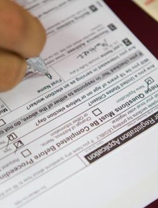 A Texas Voter registration application lays on a table; we can see a brown hand with a pen as someone fills it out. A Texas driver's license is off to one side.