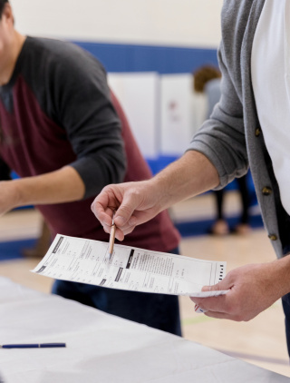 A person in a white shirt and unbuttoned grey cardigan uses a pen to point at a form in a polling place. They are standing in front of a table with a white table cloth, speaking with someone else who is seated just in front of the camera.