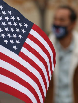 A handheld American flag is held in front of the camera. Slightly blurred in the background, we can see a man in a brown jacket with a white collar who is wearing a mask over his nose and mouth.