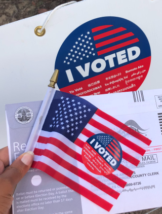 A close-up photo of a brown hand holding an American flag, a red white and blue "I voted" sticker, and a mail ballot envelope.