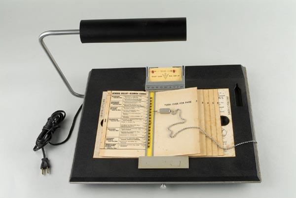 Image of a punch-card voting machine featuring a sample ballot in a punch-card holder. The stylus is attached to the machine via a chain. Image provided by UC Berkeley.