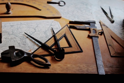 Mathematical tools, including a compass, a protractor, and a compass, lie on a wooden table on top of paper maps.