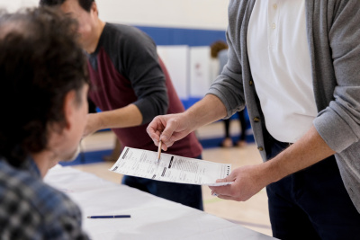 A person in a white shirt and unbuttoned grey cardigan uses a pen to point at a form in a polling place. They are standing in front of a table with a white table cloth, speaking with someone else who is seated just in front of the camera.
